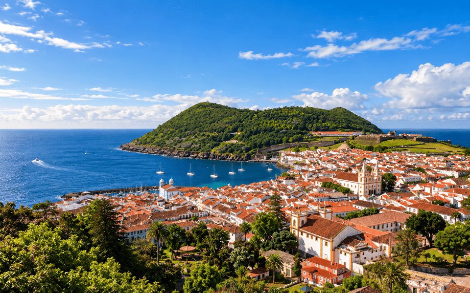 High-angle view of Angra do Heroísmo on Terceira in the Azores, with white buildings, orange rooftops, a blue harbour and the green Monte Brasil peninsula