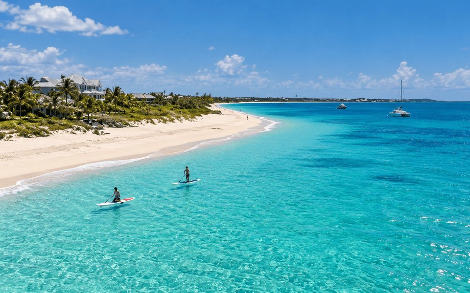 Paddleboarders in clear turquoise water off a white sand beach in Providenciales for November flight deals from Montreal